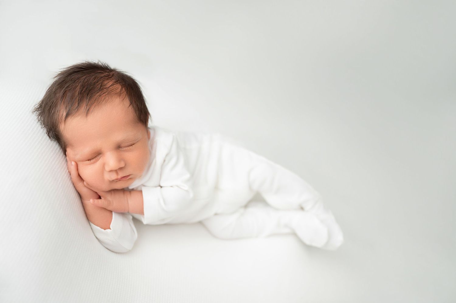 Sleeping newborn baby wearing white clothing curled up on soft white backdrop.