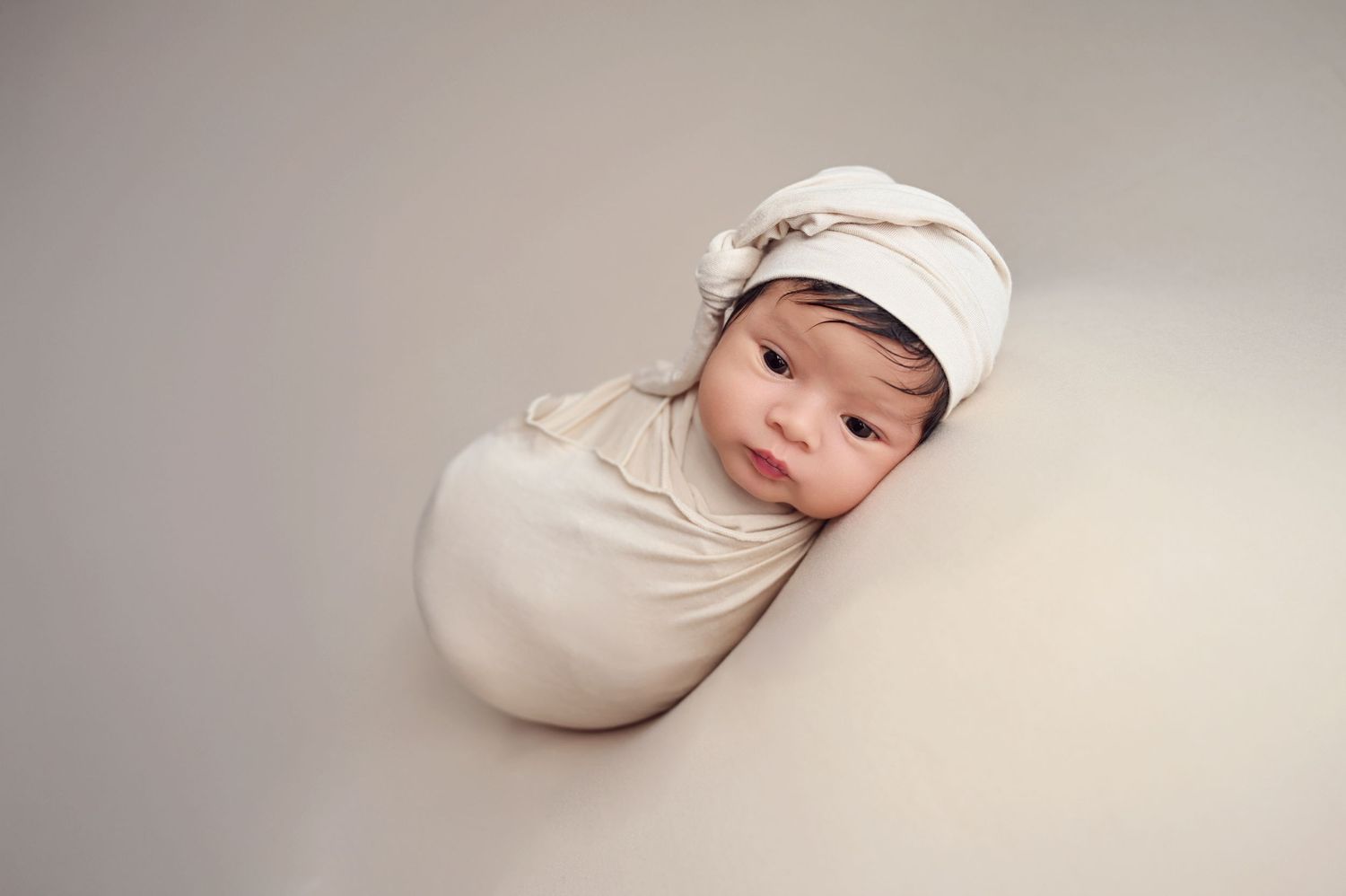 Newborn baby swaddled in cream-colored wrap and matching cap sleeping peacefully against neutral backdrop. Fresno Newborn Photography