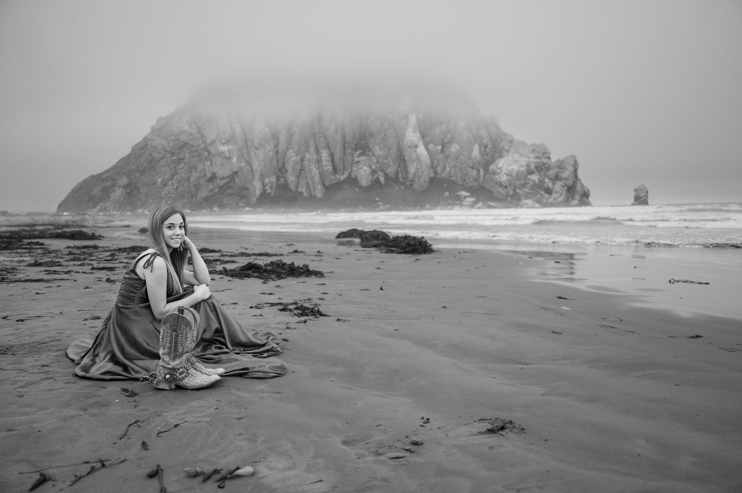 A person sits on a misty beach with dramatic rocky cliffs in the background creating a moody coastal scene.