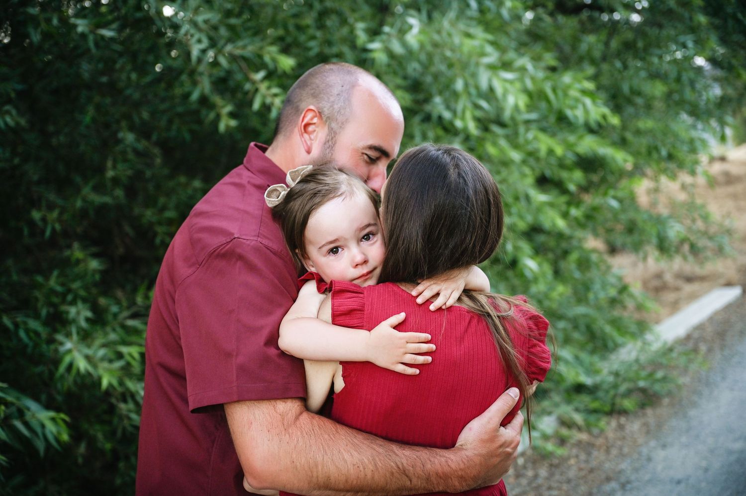 A family shares a warm embrace outdoors on a path with green foliage in the background. Hanford Family Photographer