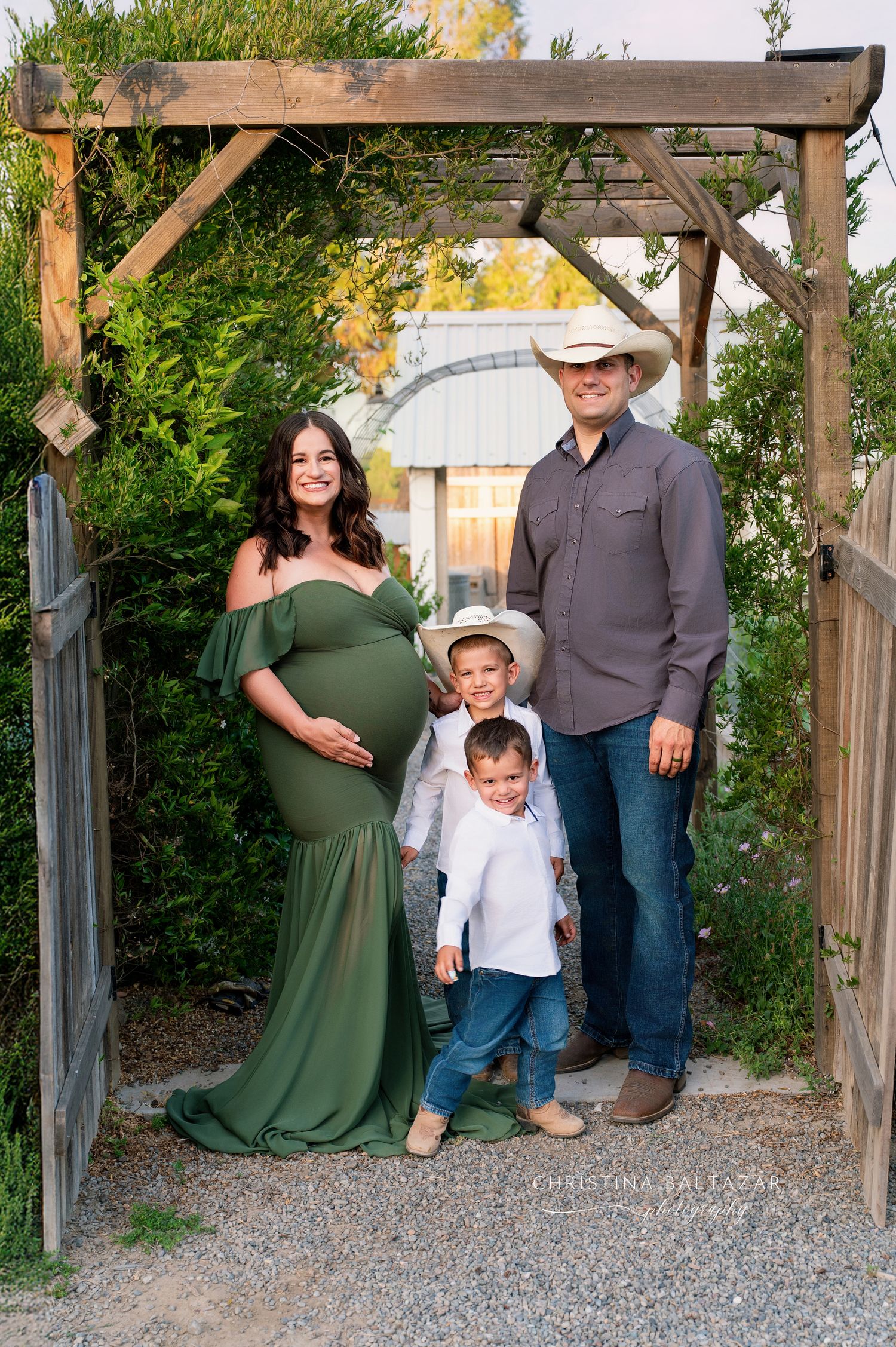 A family poses together under a rustic wooden archway in a garden setting, wearing coordinated casual attire. Fresno Maternity Photography