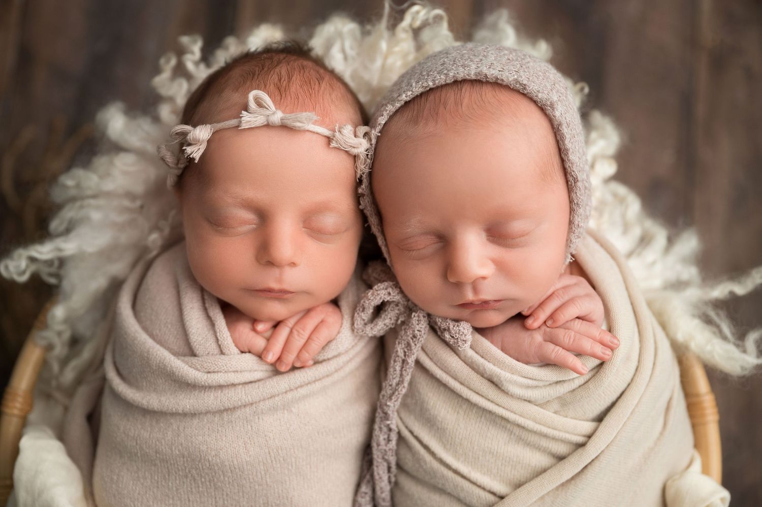 Sleeping twin newborns wearing matching white bonnets on soft cream textured backdrop.