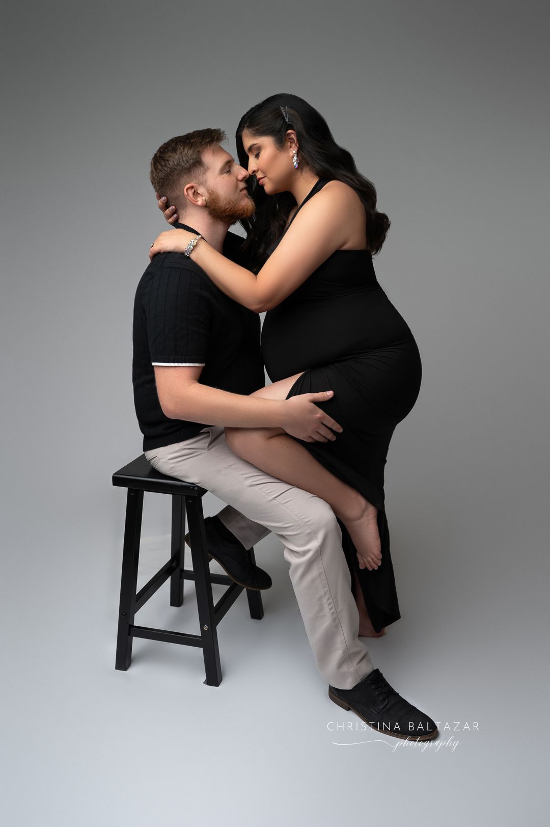 Intimate maternity portrait of couple embracing on wooden stool against gray backdrop in studio setting. Fresno Maternity Photographer