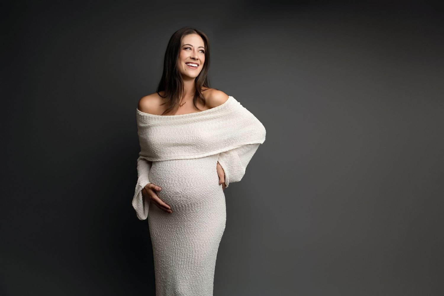Smiling pregnant woman in elegant off-shoulder white knit dress posing against dark gray background in studio portraits in Lemoore.
