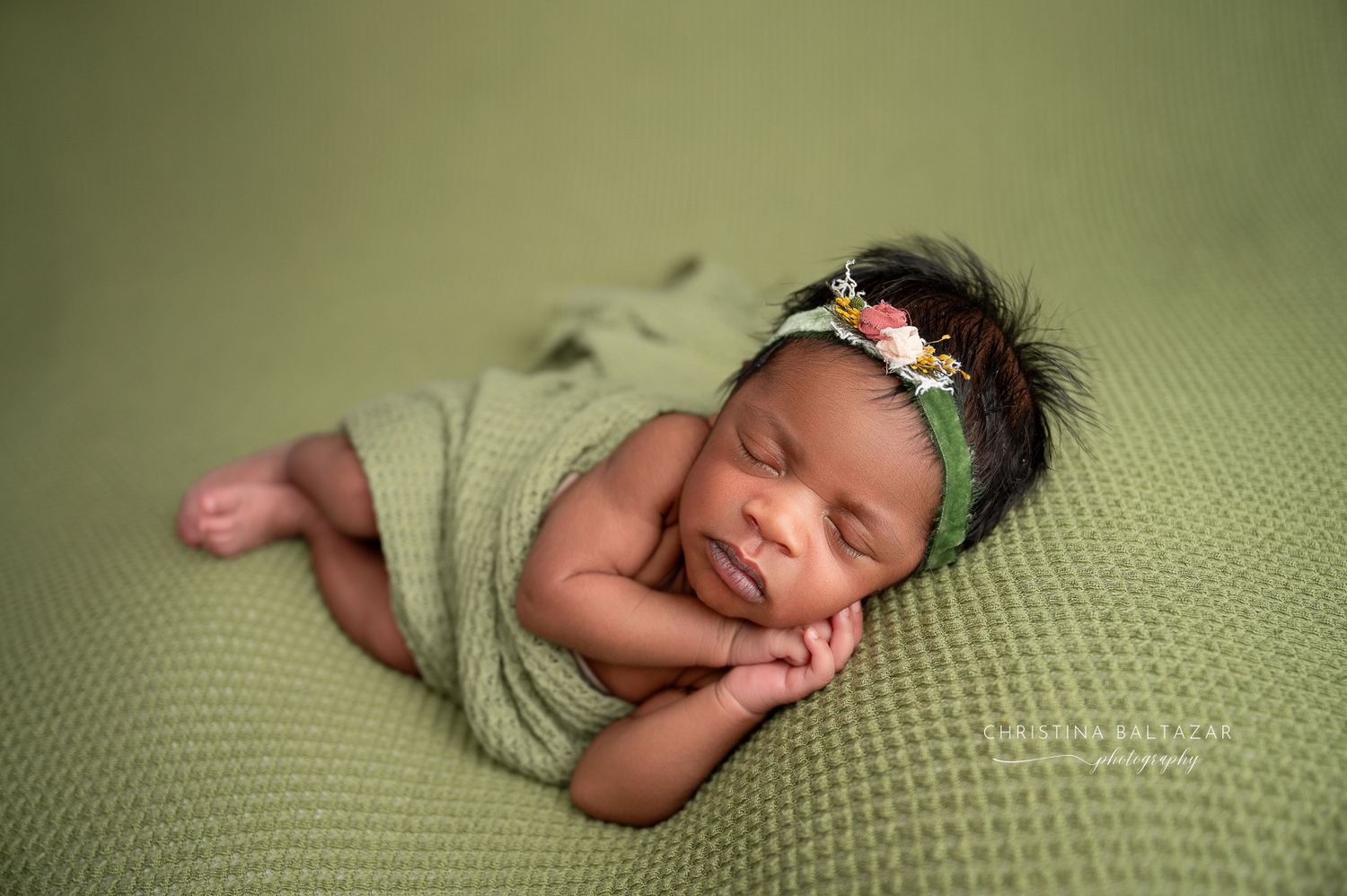 Newborn baby sleeping peacefully on green blanket while wrapped in soft swaddle with floral headband. Fresno Newborn Photographer