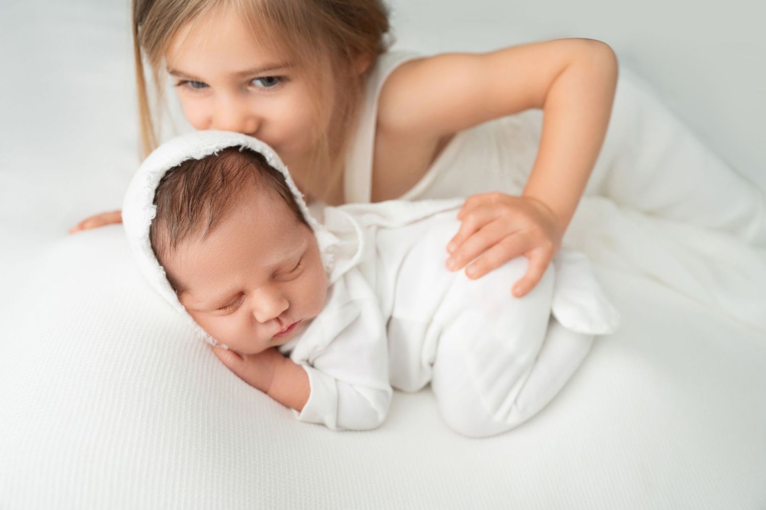 A sleeping newborn baby in white clothing rests on soft white bedding while being tenderly cradled. Fresno Newborn Photographer