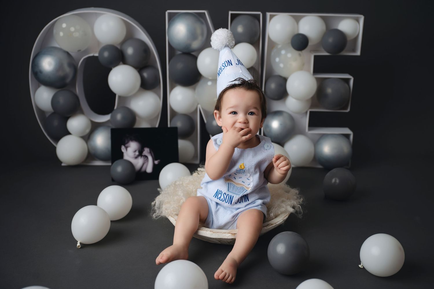 Baby milestone photoshoot with black and white balloons against dark background.