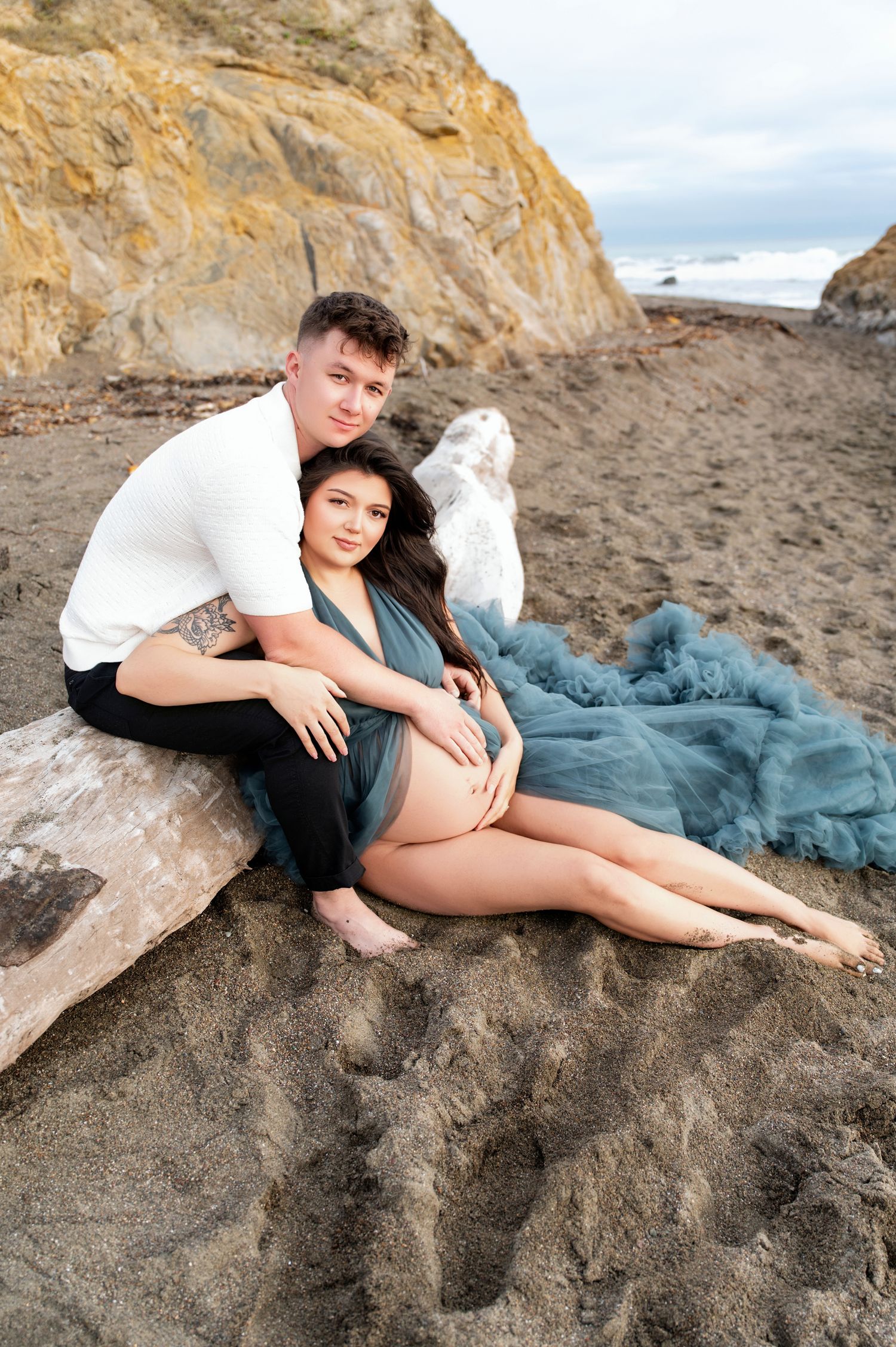 Maternity beach photo with couple sitting on driftwood in teal gown.