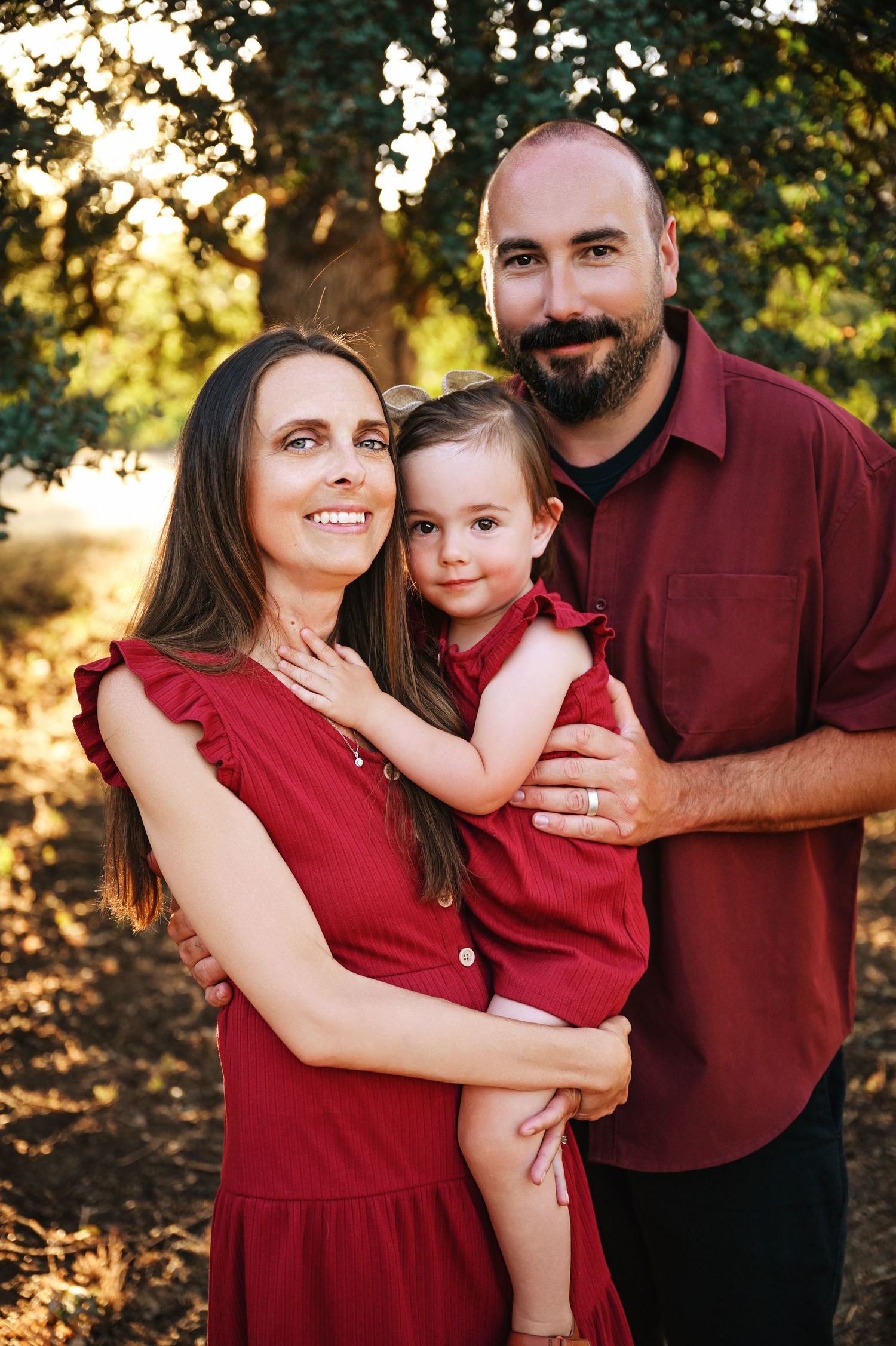 Family wearing coordinated red outfits poses together outdoors during golden hour sunset with trees in background. Visalia Family Photograph