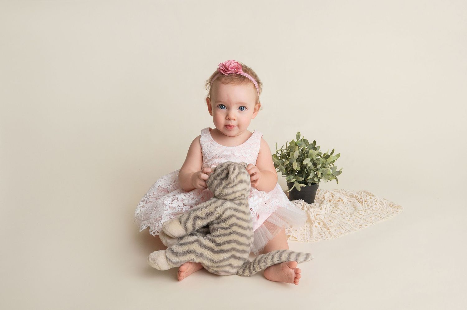 Baby in white outfit sits on floor with decorative elements during photo session.