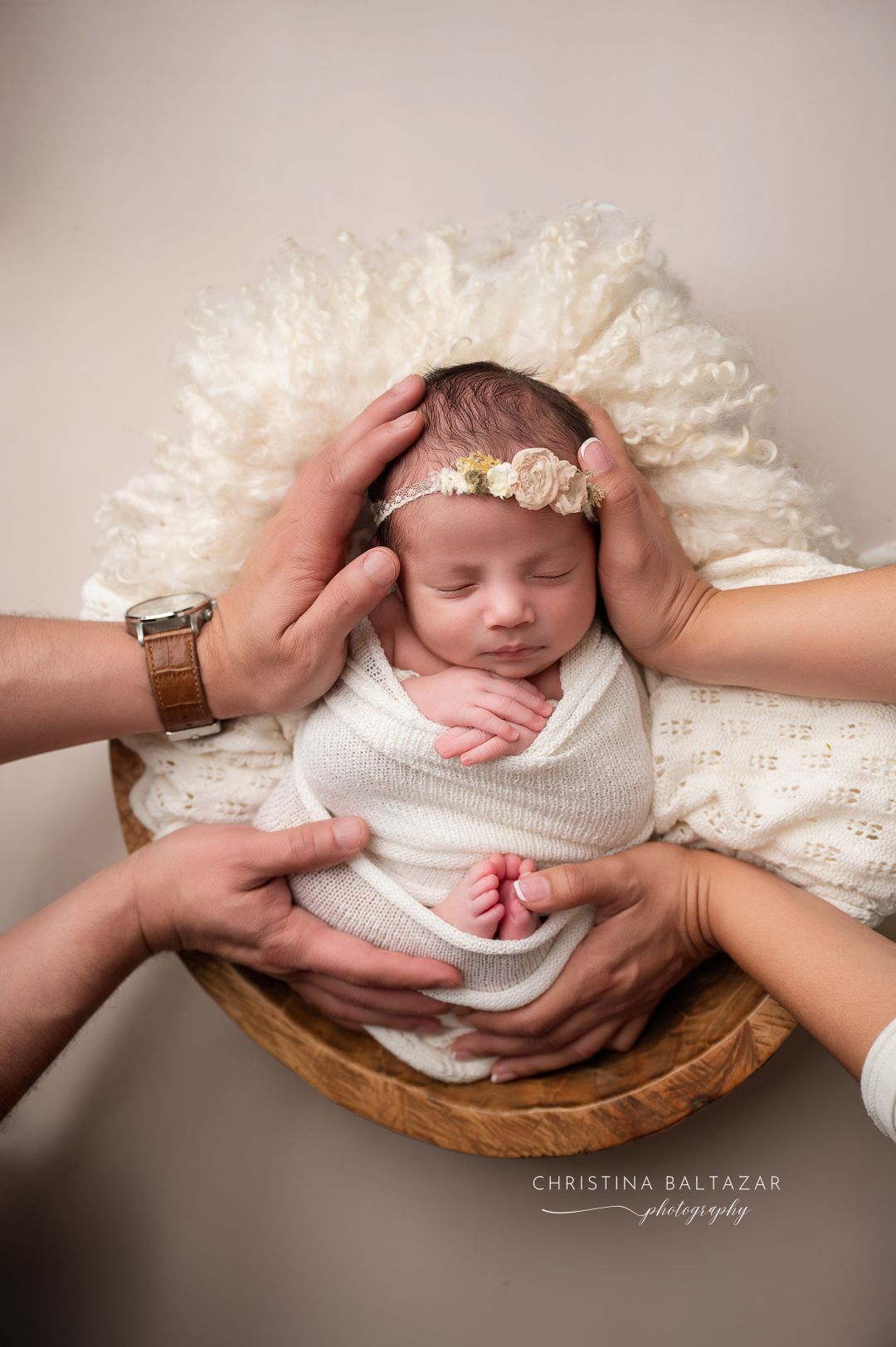 A sleeping newborn being gently supported by adult hands while lying in a wooden bowl on white fluffy blankets. Fresno Newborn Photographer