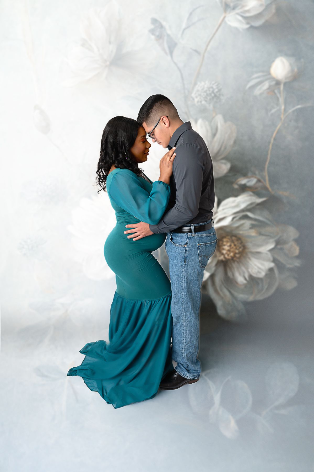 Couple sharing a tender moment, woman in teal maternity gown, man in gray shirt and jeans, floral studio backdrop. Fresno maternity photos