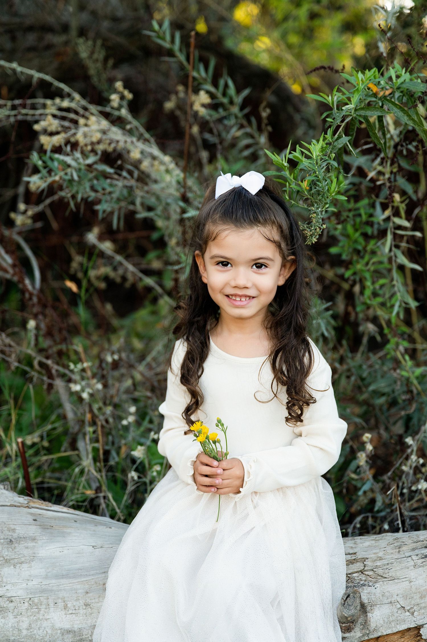 A child in a white dress sits outdoors among greenery and flowers on a sunny day. Fresno Family Photography