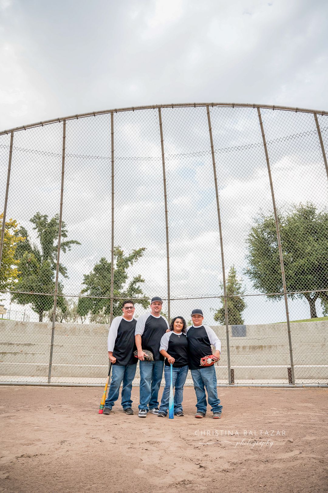 Four individuals in matching black shirts stand on a baseball field in front of a tall chain-link backstop. Hanford Family Photographer