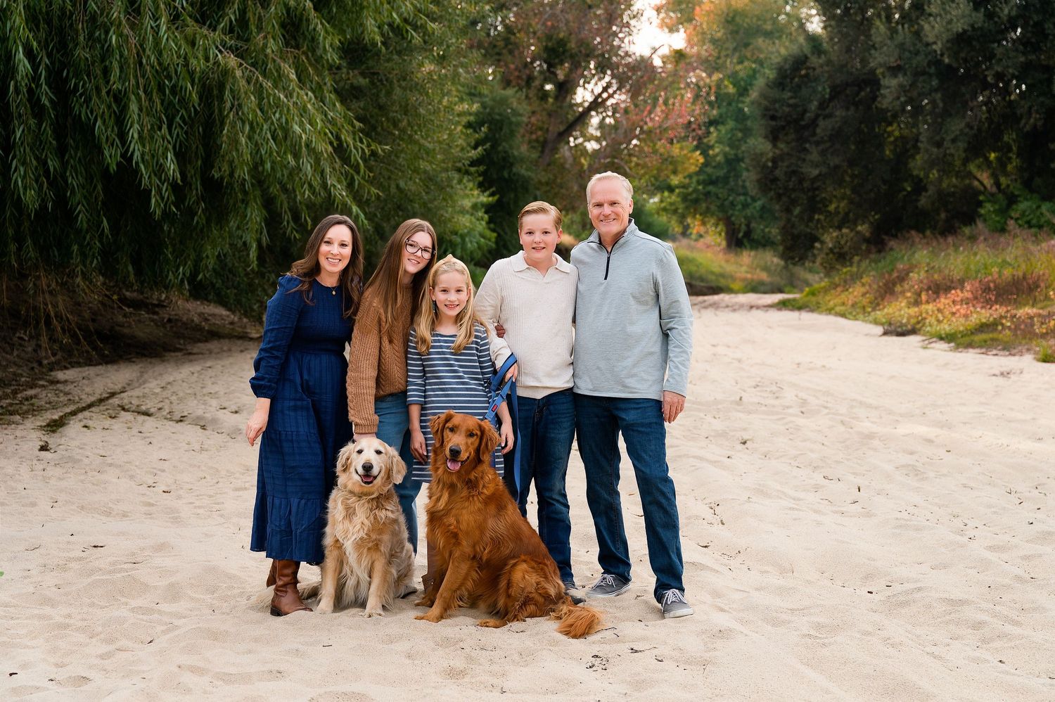 Family and pets pose together for outdoor portrait session on sandy beach path.