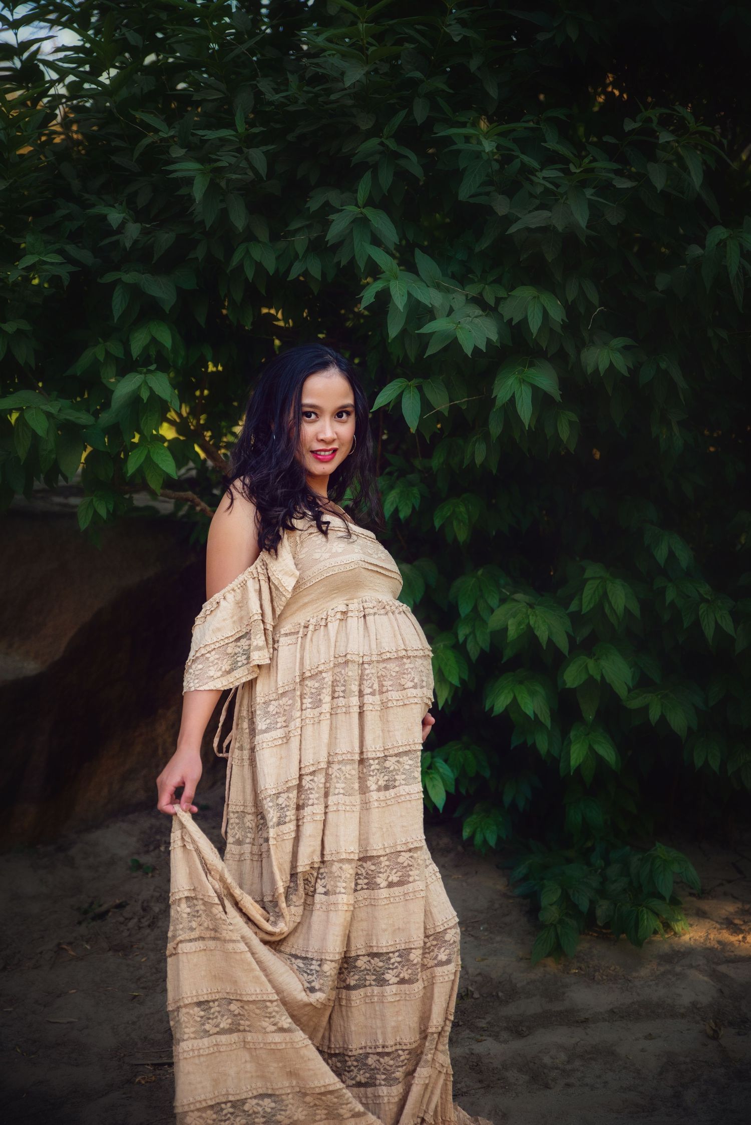 A pregnant woman in a beige dress poses outdoors against a backdrop of green foliage, in Visalia, CA.