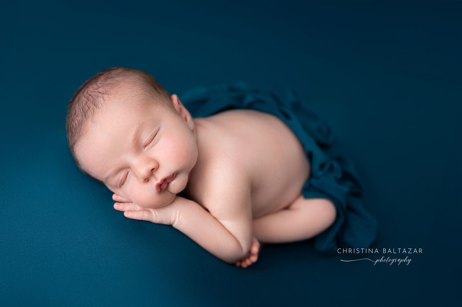 Sleeping newborn baby curled up peacefully on deep blue backdrop in professional portrait studio setting. Fresno Newborn Photographer
