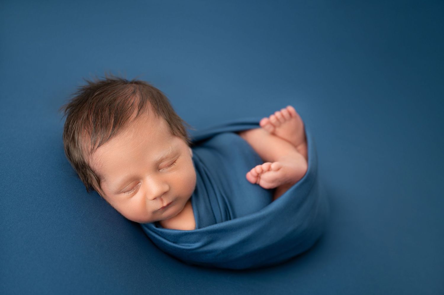 Smiling newborn wrapped in blue fabric against deep blue background.