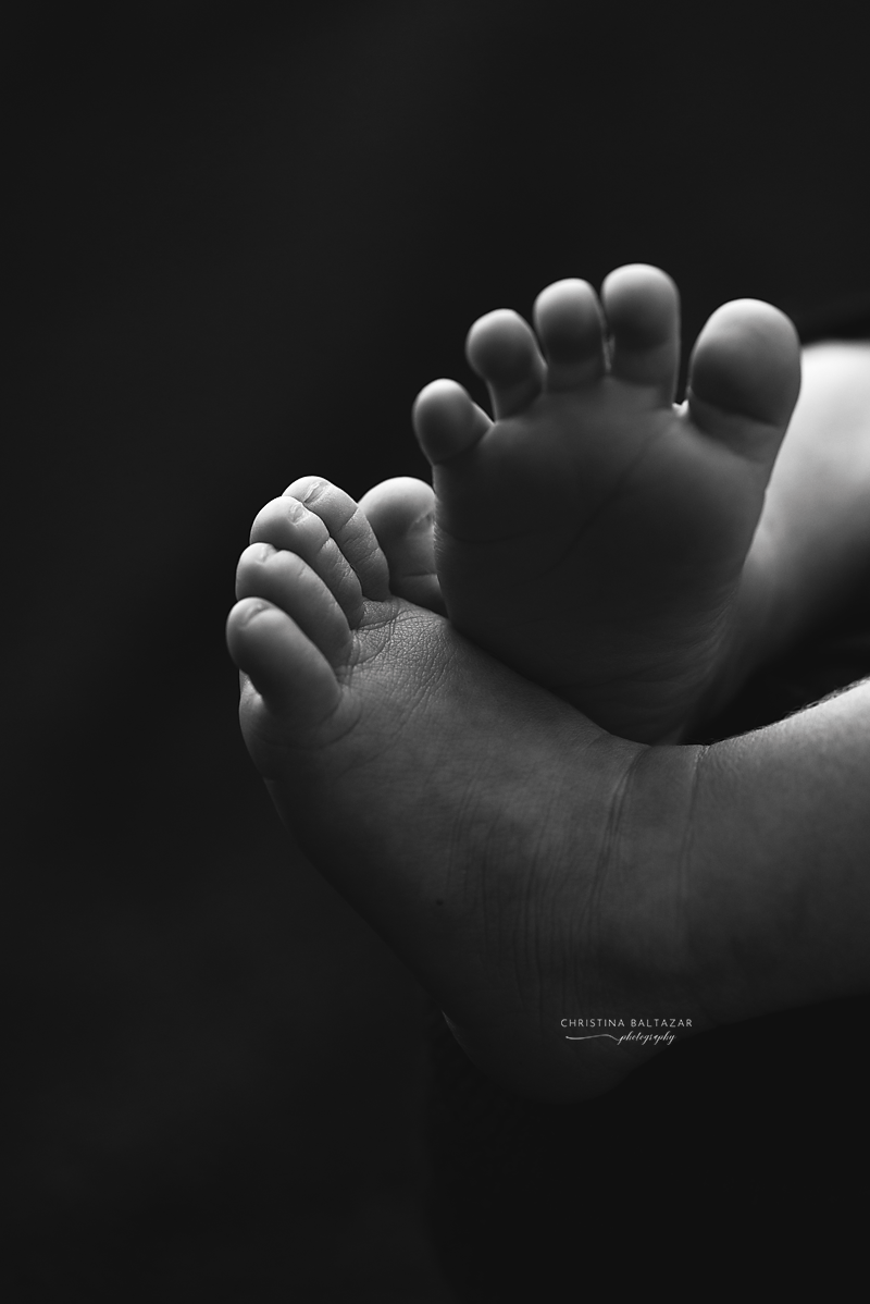 Dramatic black and white photograph of tiny baby feet with delicate toes against dark background.