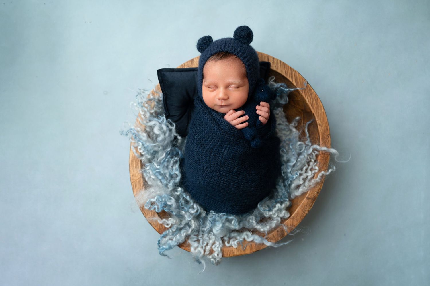 Newborn in dark blue knit wrap and bear bonnet sleeps peacefully in round wooden bowl.