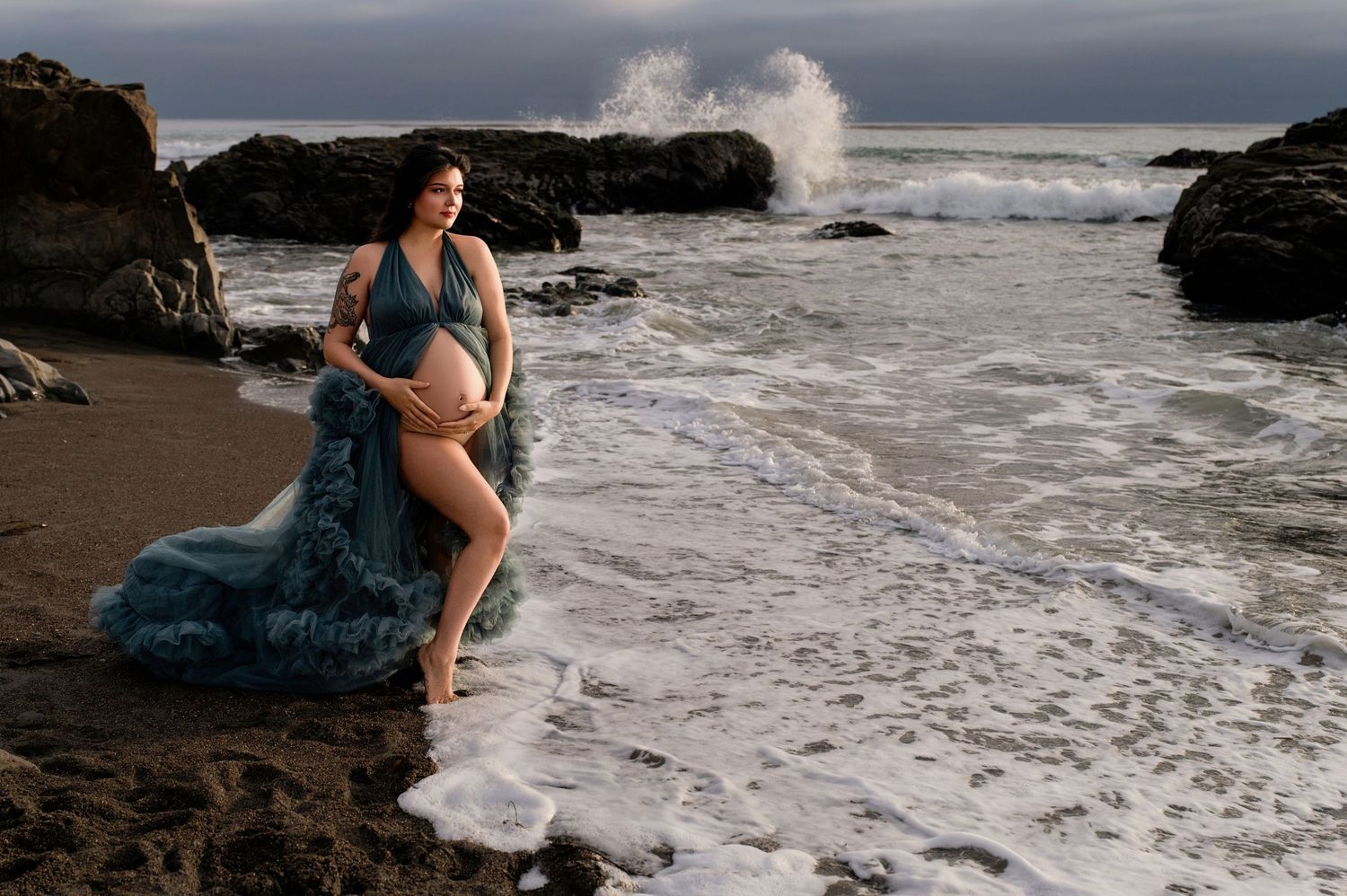 Romantic beach photoshoot at sunset with waves crashing against dark rocks in the background.