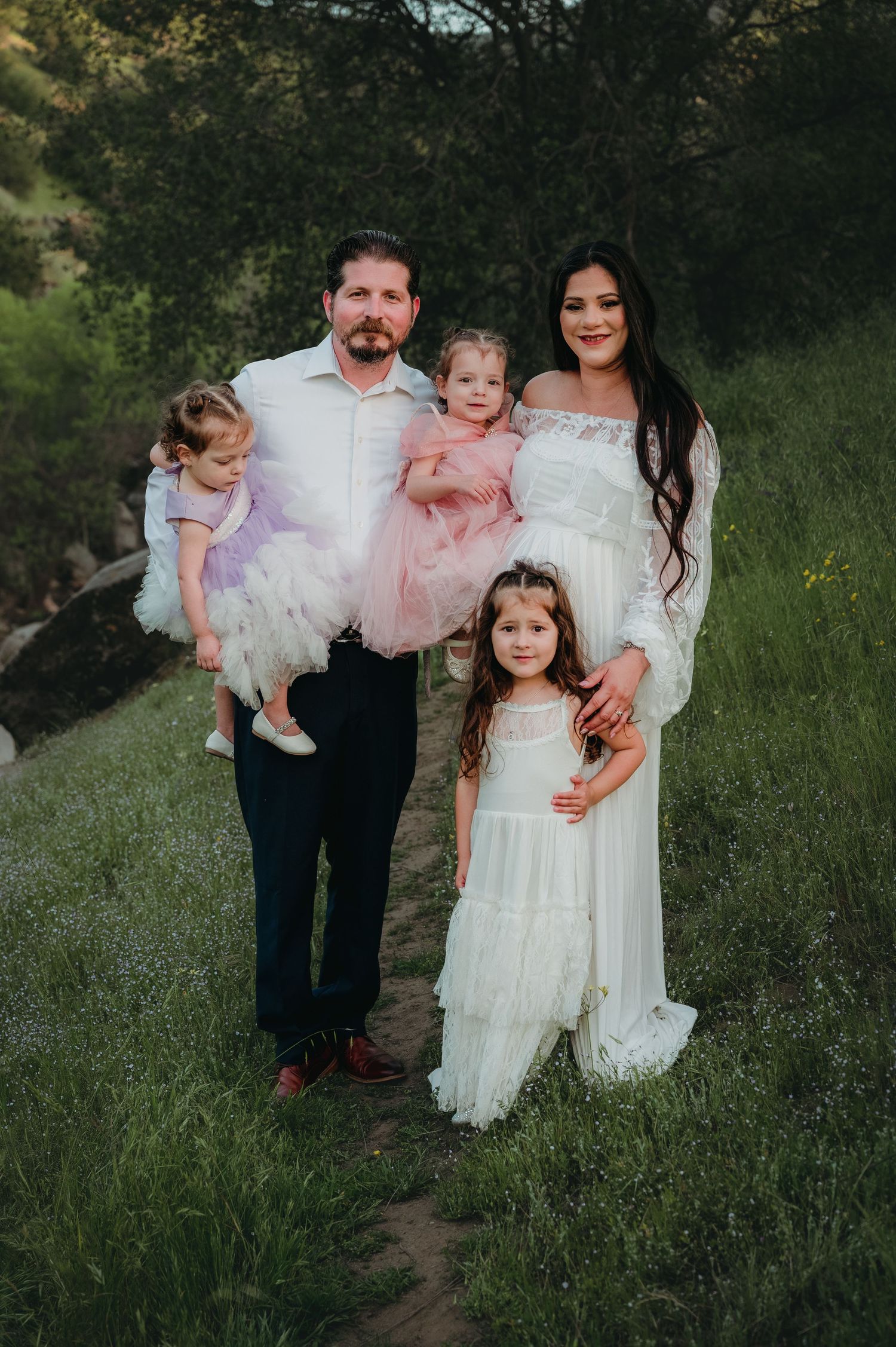 Family in coordinating white and pink outfits poses together on a rustic outdoor path. Fresno Family Photography