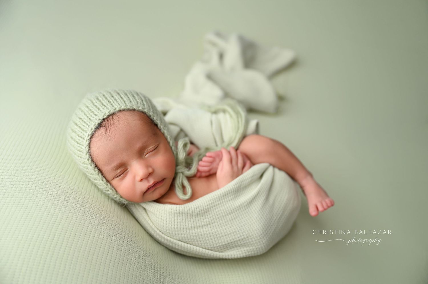 Newborn baby sleeping peacefully in soft white swaddle blanket on light green background. Fresno Newborn Photography