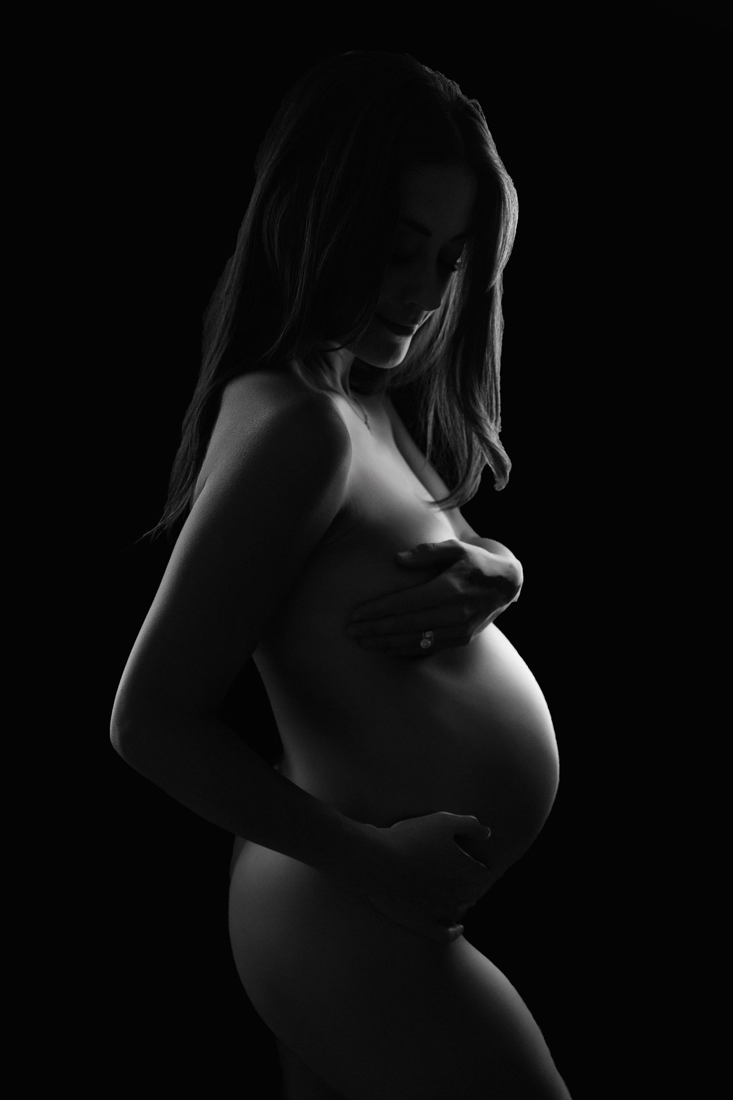 Artistic black and white silhouette of pregnant woman holding baby bump against dark background in studio maternity shoot in Fresno, CA.