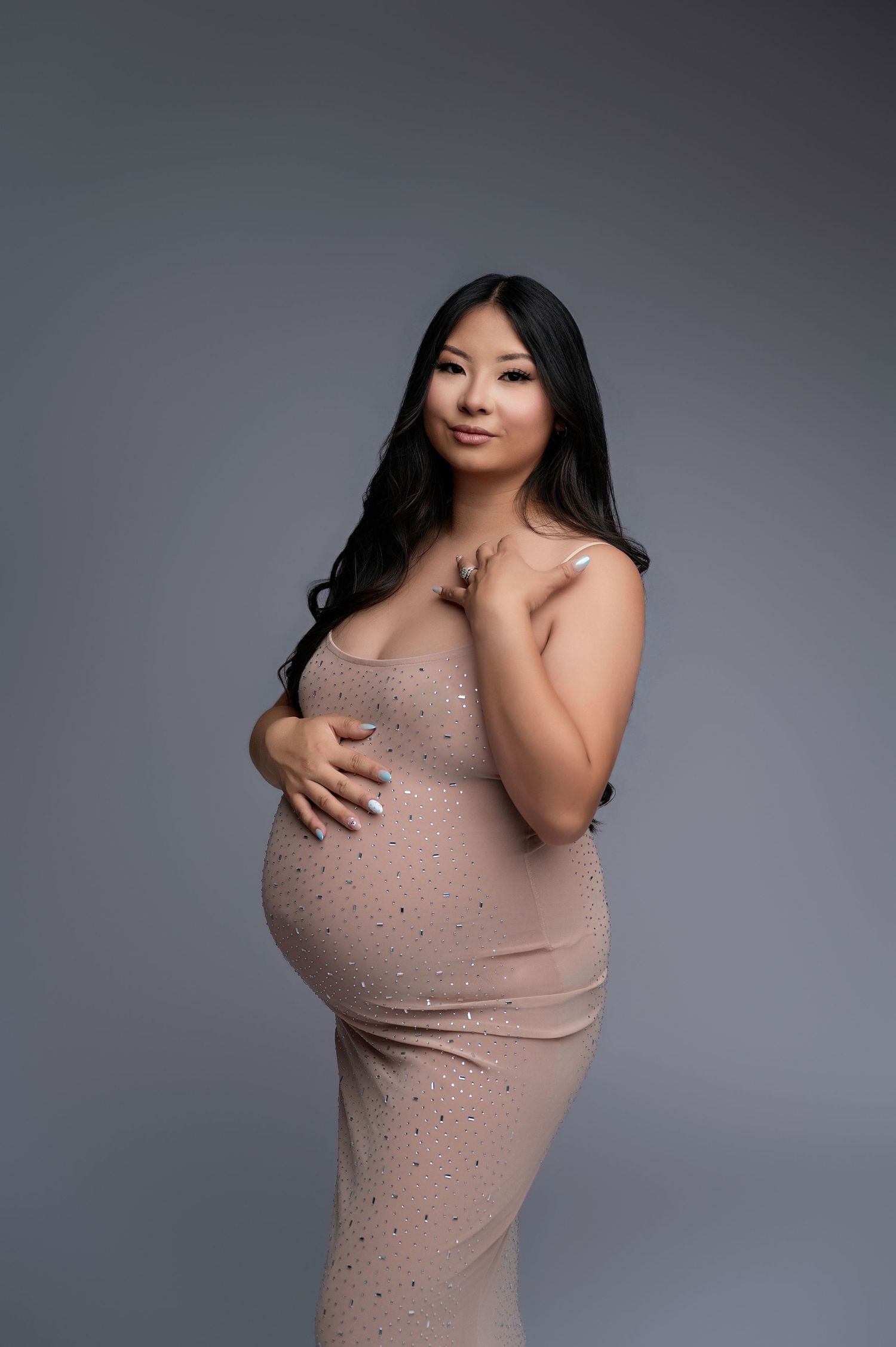 Pregnant woman in a beige rhinestone dress cradling her baby bump in a professional studio maternity photo shoot.