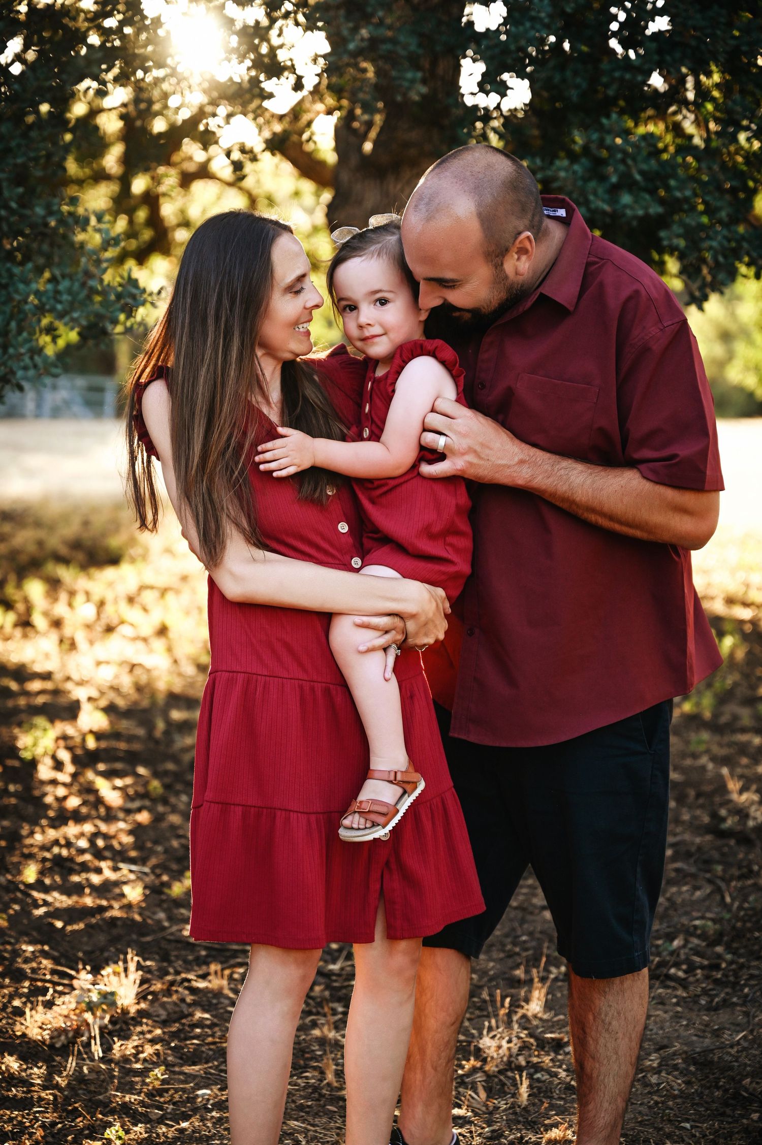 A family wearing matching burgundy outfits poses together outdoors during golden hour in a natural setting. Visalia Family Photographer