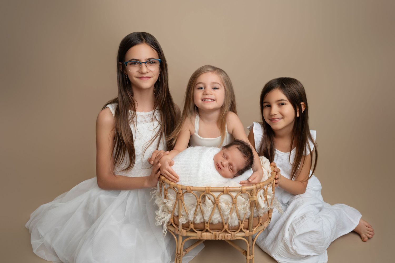 Three girls in white dresses pose with sleeping baby in wicker basket against beige backdrop.
