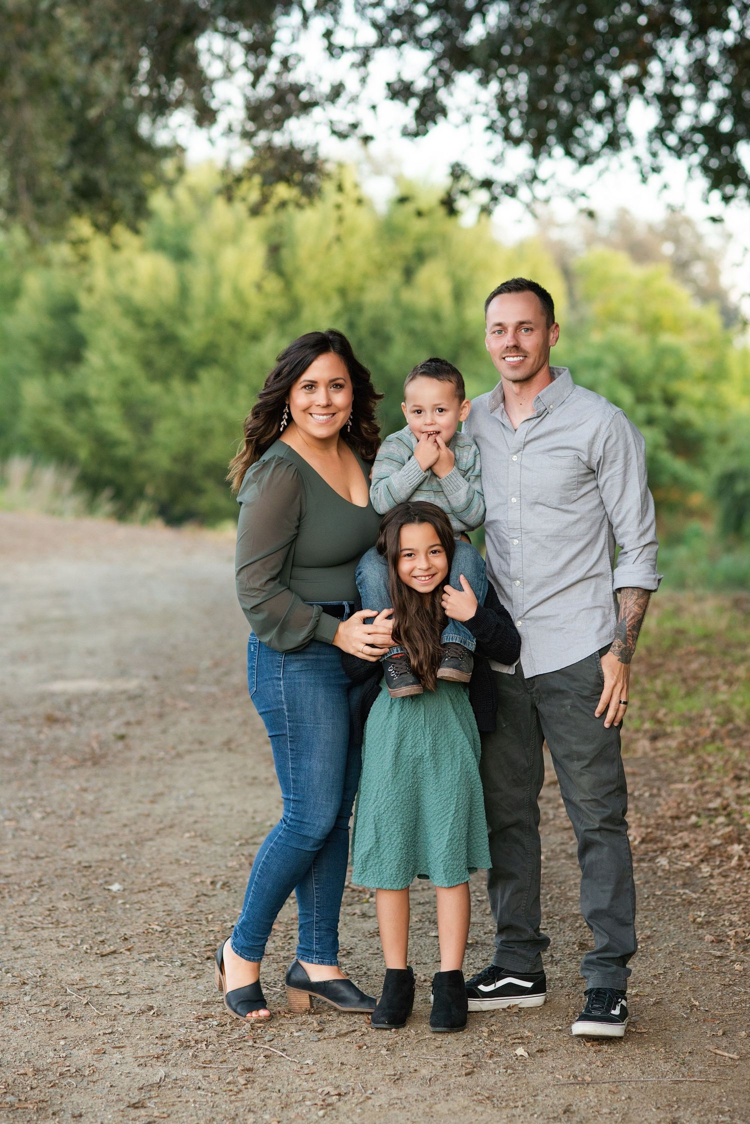 A family shares playful moments together on a dirt path surrounded by green foliage during an outdoor photo session. Hanford Family Photos