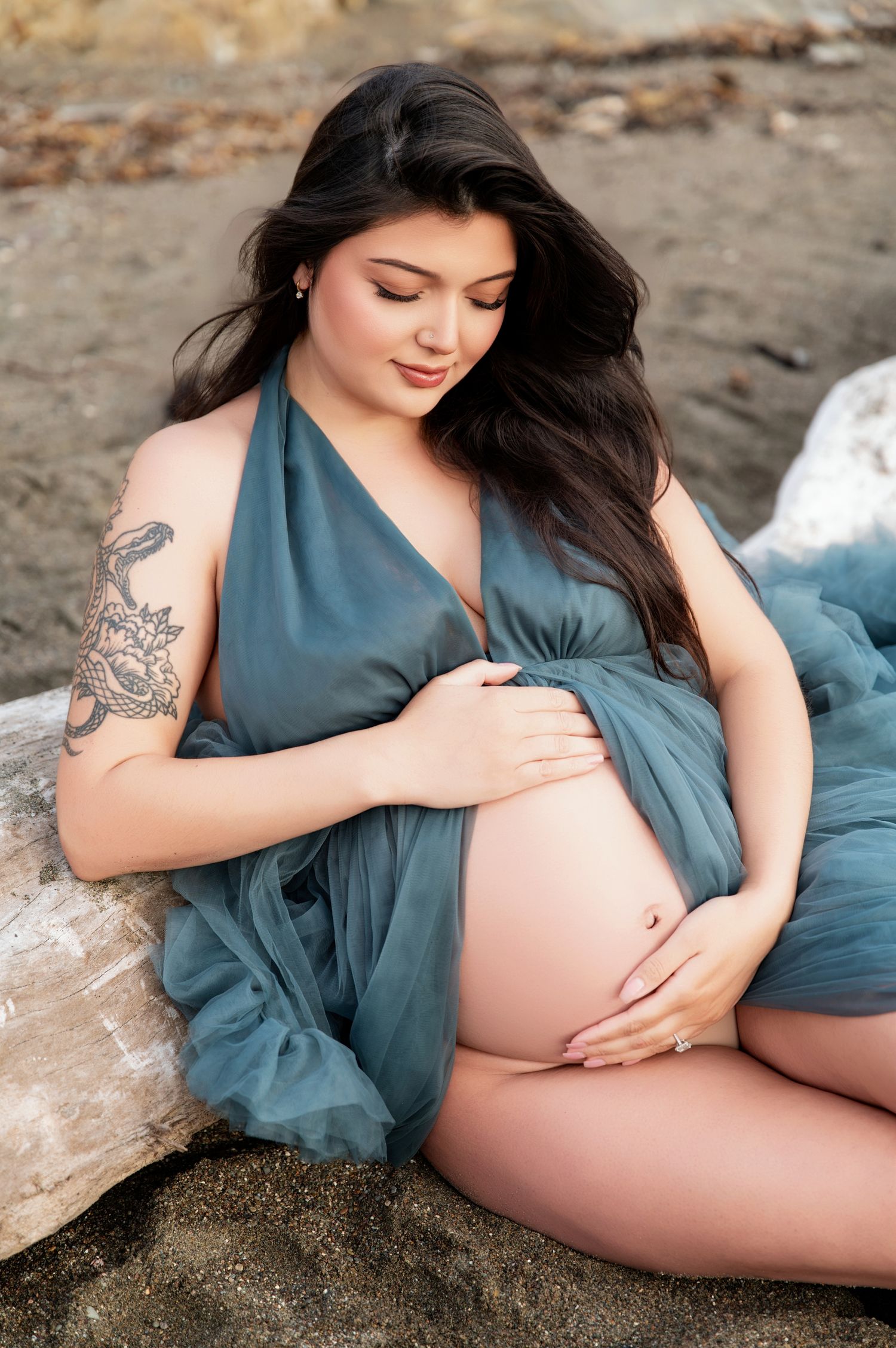 Expectant mother in teal dress poses on beach rocks showing her baby bump.