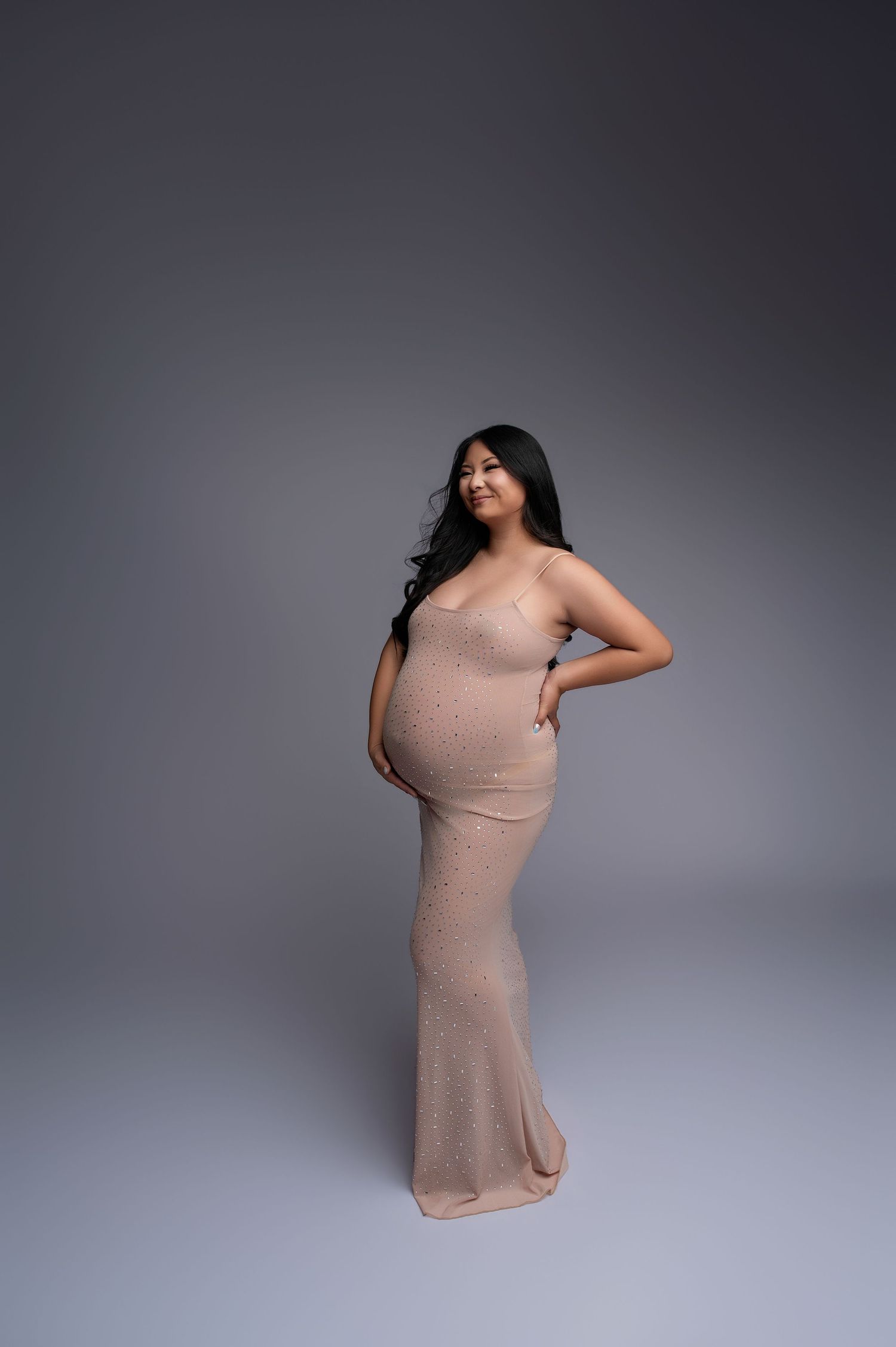 Pregnant woman in a form-fitting rose gold glitter gown posing elegantly against a gray studio backdrop.