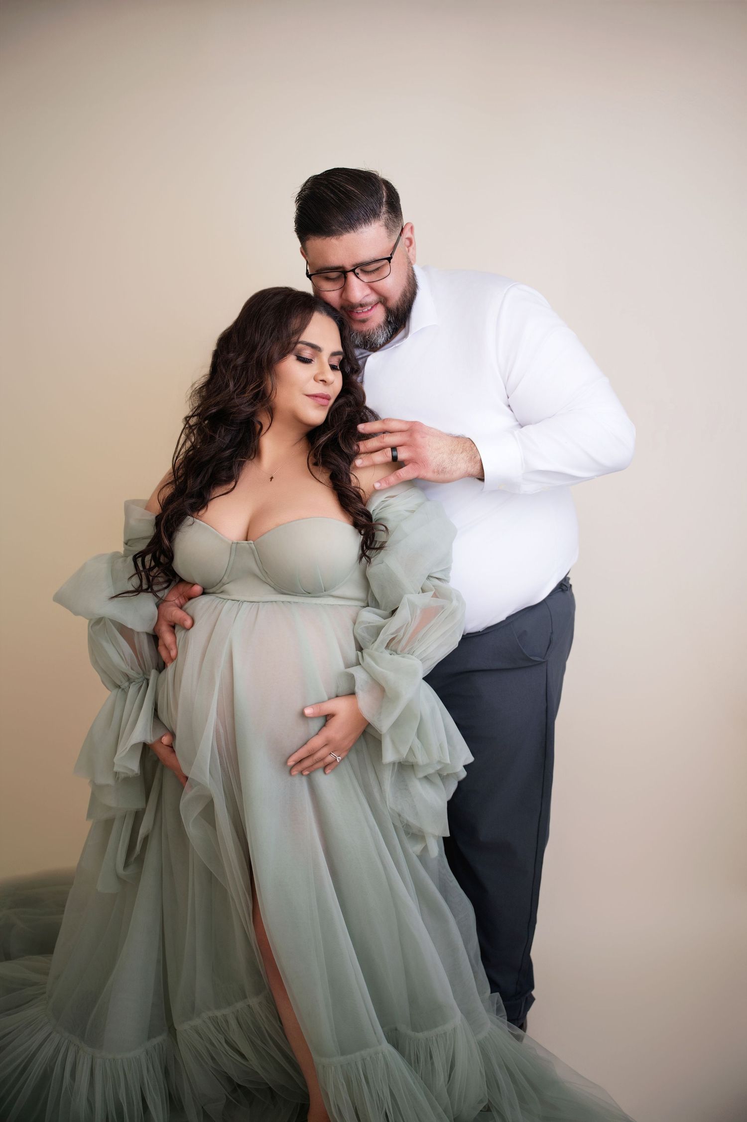 Romantic maternity photos with couple in sage green gown against neutral backdrop.