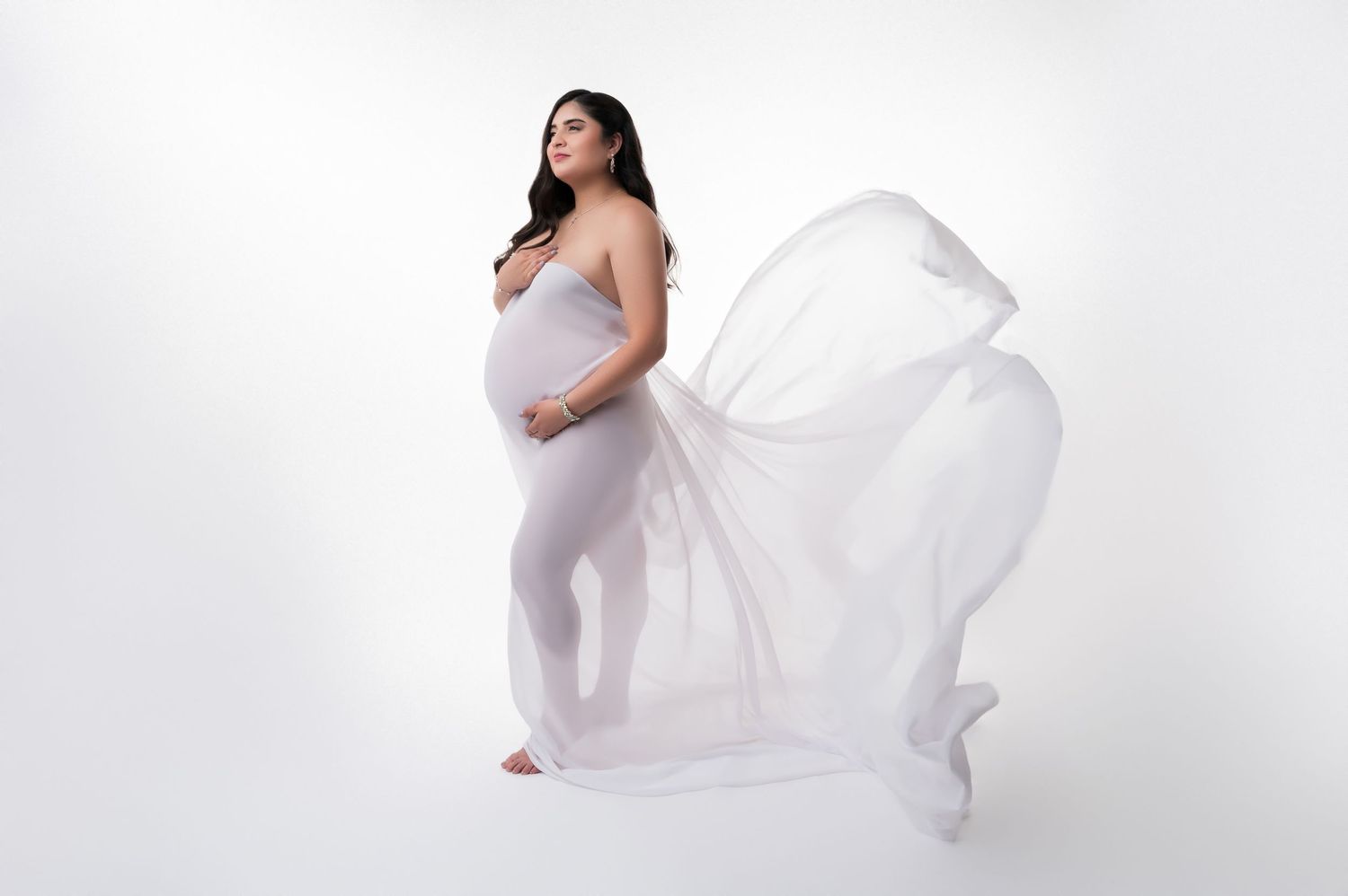 Pregnant woman in white flowing fabric draped elegantly, showcasing her baby bump against a white background. Fresno Maternity Photos