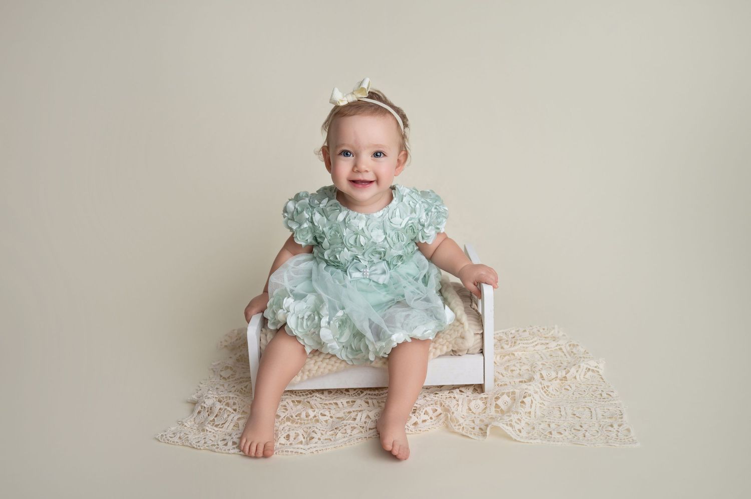 Sweet baby in seafoam dress sits on lace-covered floor during photo session.