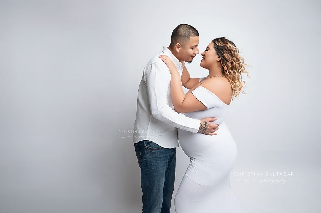 A couple embraces tenderly in a maternity photo shoot against a light gray studio background. Hanford Maternity Photographer