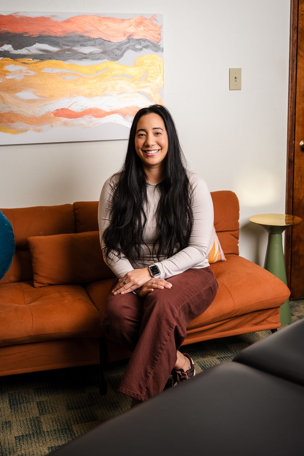 Smiling woman with long black hair sits on an orange sofa in a modern room with colorful abstract wall art.