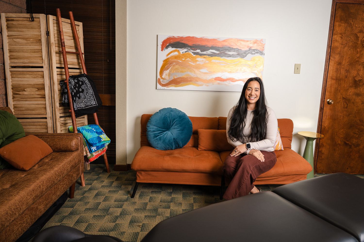Woman sitting on orange sofa in cozy room with colorful abstract painting, teal pillow, and wooden decor.