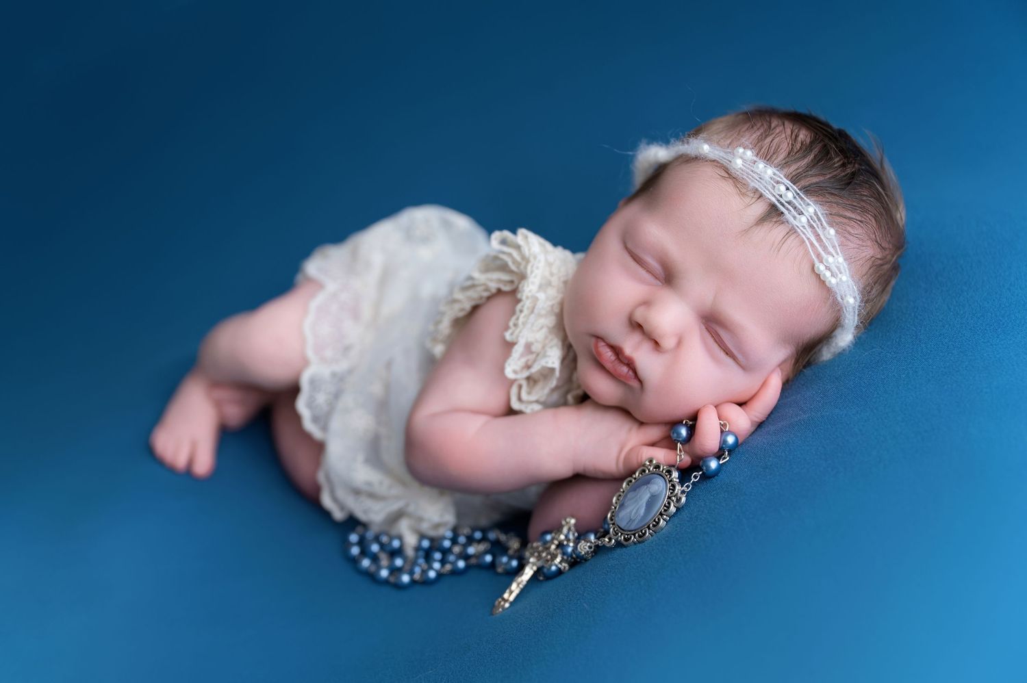Fresno Newborn Sleeping newborn baby in white lace outfit with pearl headband resting on blue backdrop with decorative brooch.