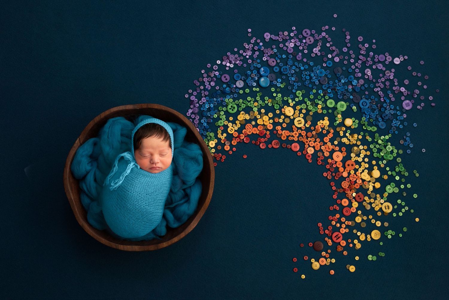 A newborn portrait of a baby, wrapped in blue, with a rainbow of buttons on a dark blue backdrop.