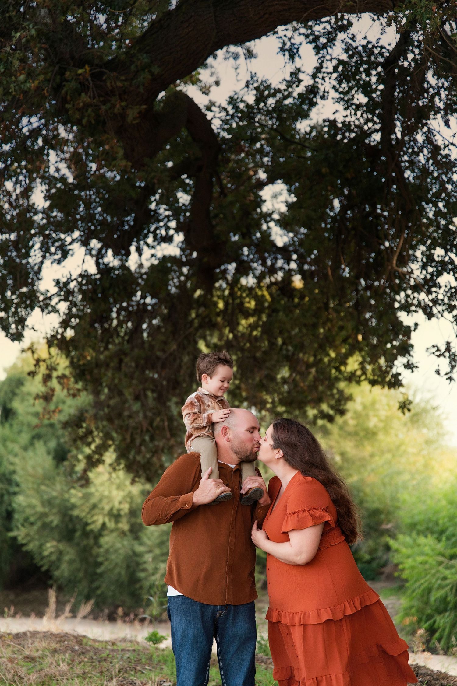 Couple in coordinating rust-colored outfits share intimate moments under a large oak tree. Hanford Family Photographer