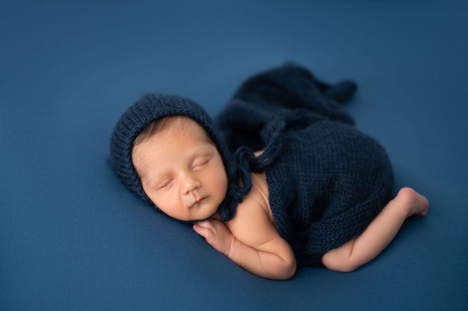 Sleeping newborn wrapped in navy knit wrap on deep blue background.