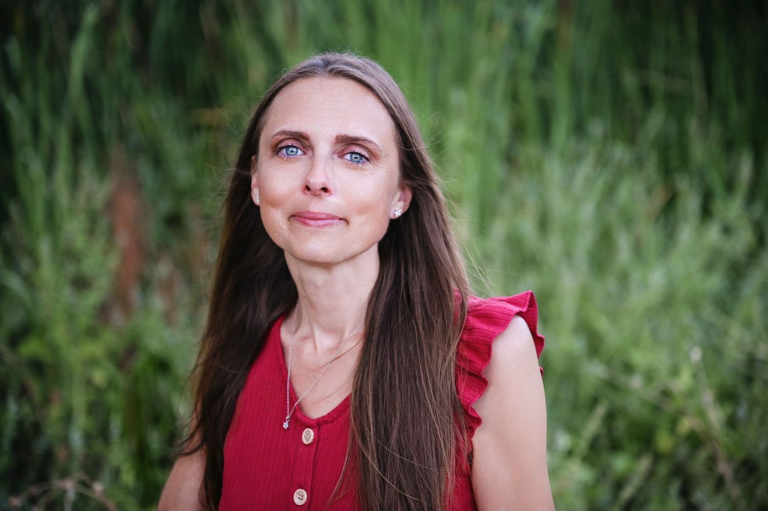 A woman in a red blouse smiles joyfully outdoors with a lush green background. Visalia Family Photographer