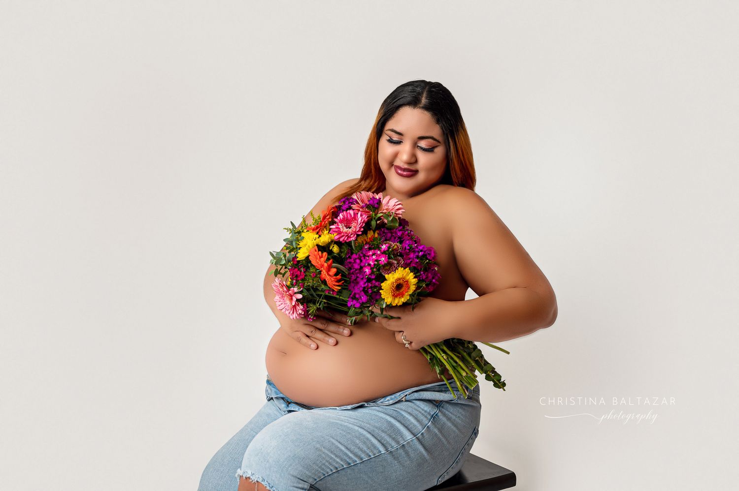 A pregnant woman in light blue jeans holds a vibrant bouquet of pink and yellow flowers against a white backdrop. Fresno Maternity Photos