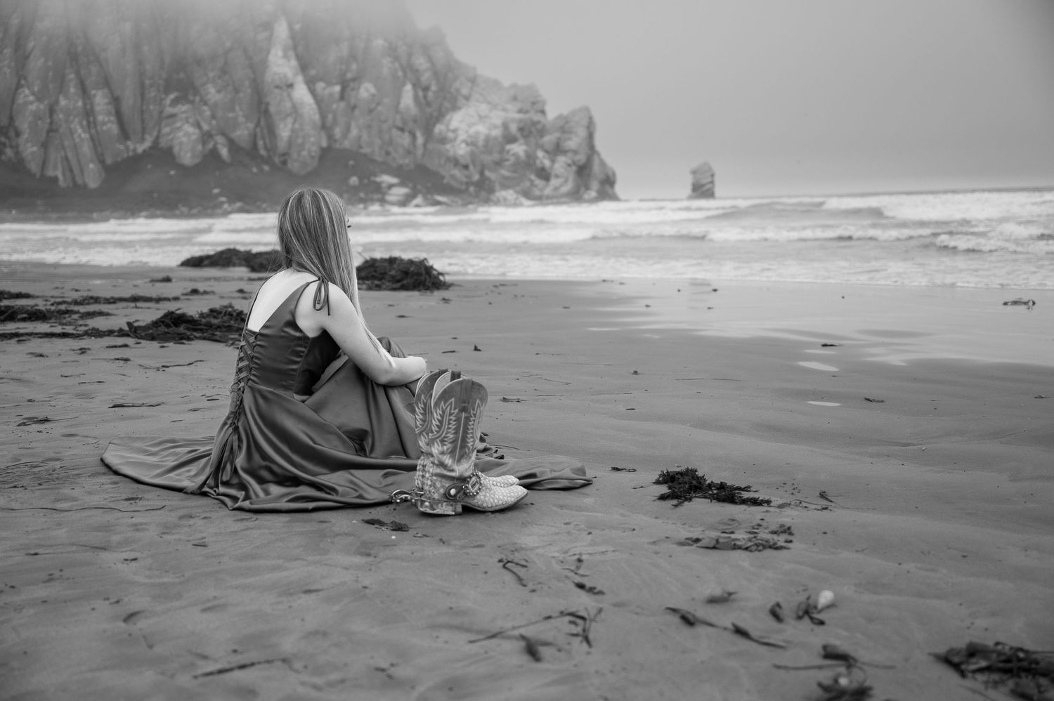 A black and white image shows a solitary figure sitting on a moody beach with rocky cliffs.