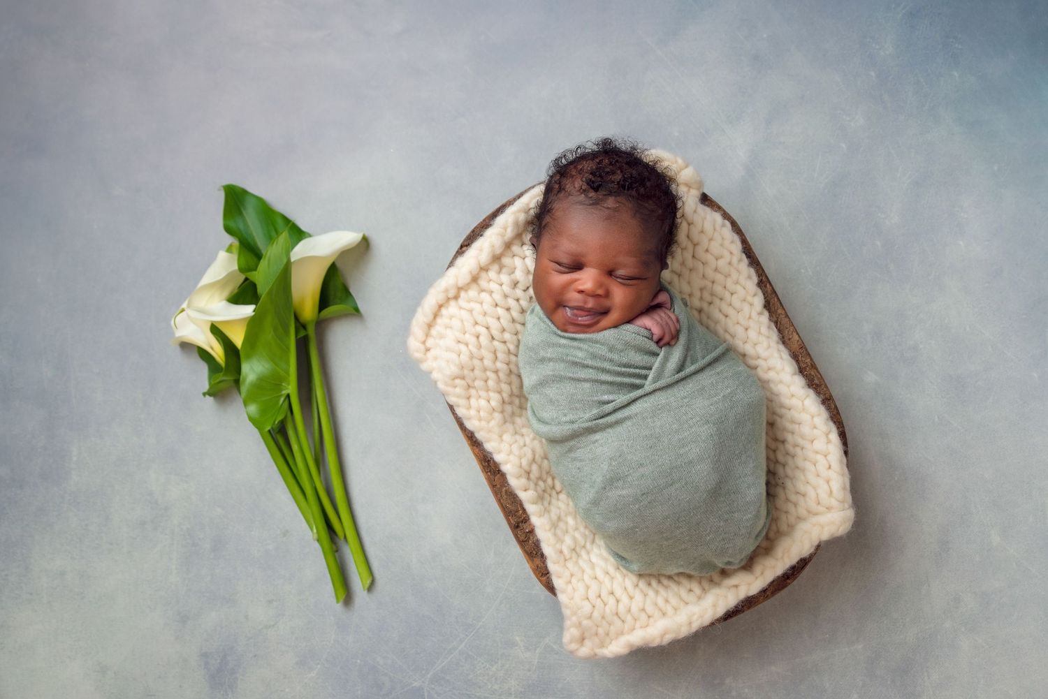 Sleeping newborn wrapped in soft mint green swaddle lying in knitted basket with white calla lilies beside them. Fresno Newborn Photographer
