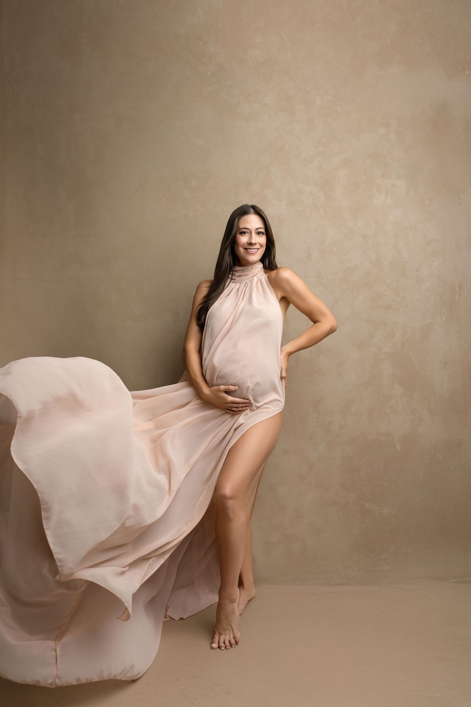 Pregnant woman in flowing blush chiffon gown smiling gracefully in a Fresno, CA studio maternity portrait.