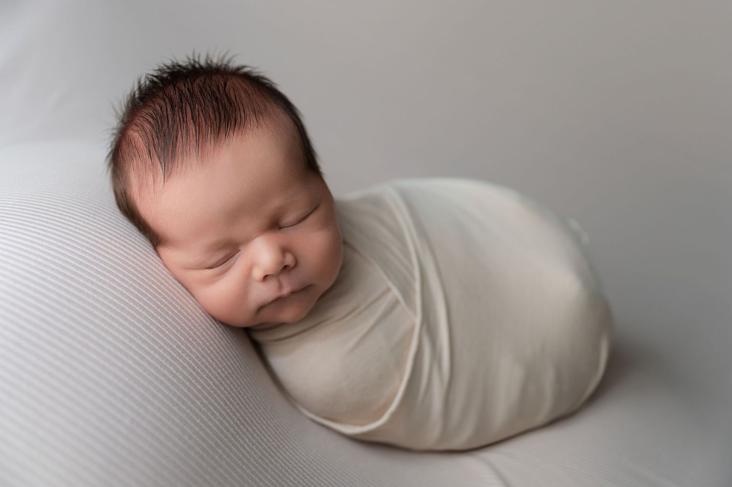 Minimalist newborn photography series with baby wrapped in cream fabric against light background.